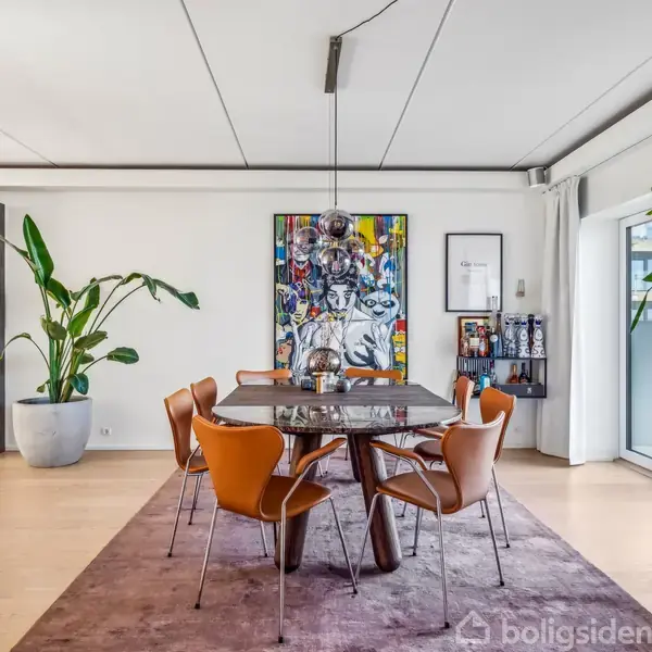 Spisebord omgivet af seks stole står på et stort tæppe. I baggrunden hænger farverige malerier, mens store grønne planter flankerer området. Dagslys strømmer ind fra store vinduer.