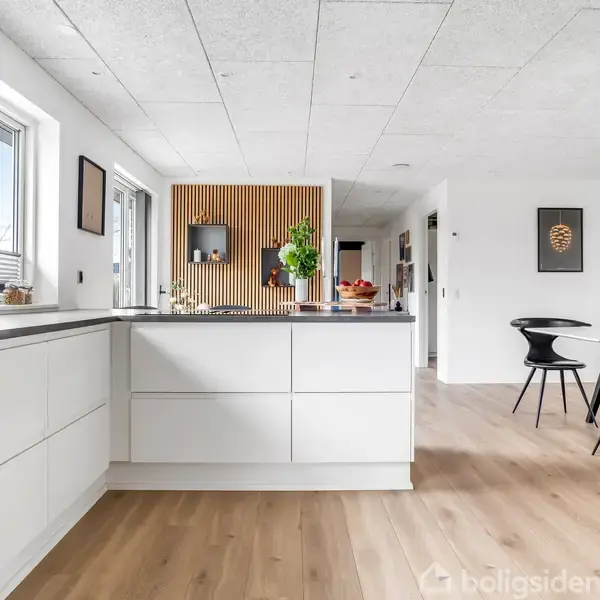 Et moderne køkken med hvide skabe, en central køkkenø med planter og frugtskål. Rummet er lyst med trægulv og tilstødende spisestue med sorte stole ved et bord.