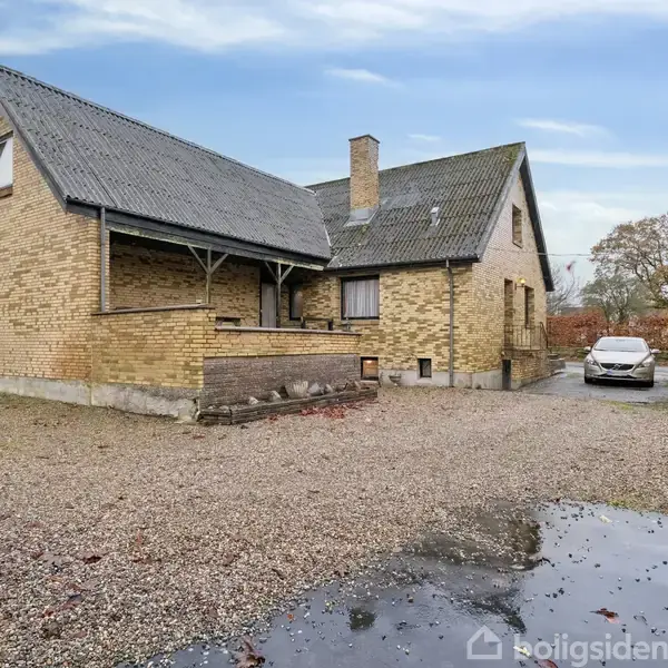 Et gult murstenshus med skråt tag står stille på en grusbelagt gårdsplads. I baggrunden ses en parkeret bil og et lille træskur omkranset af træer.