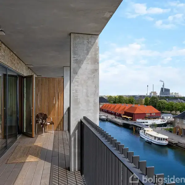 En altan med træplanker og glasdøre strækker sig langs en moderne bygning. Udsigt over en kanal, hvor både ligger til, og røde tage samt grønne områder kan ses.