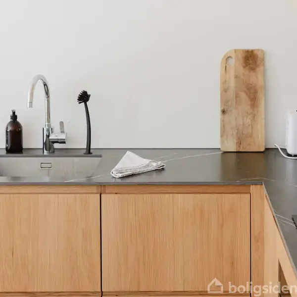 Køkkenbord med håndvask, sæbedispenser og sort børste ved siden af et skærebræt op ad væggen. Der står en brødrister længere nede på bordet.