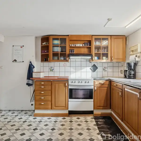 En rummelig køkken med træskabe og en central ovn står i centrum. Ved siden af er der en kaffemaskine og opvaskemaskine. Gulvet har sort-hvide fliser, omgivet af lyse vægge.