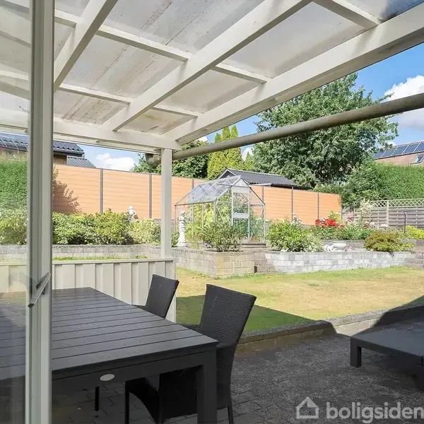 Et sort bord står under en overdækket terrasse med gennemsigtigt tag. I baggrunden ses en have med en græsplæne, et drivhus og farvede hegn.