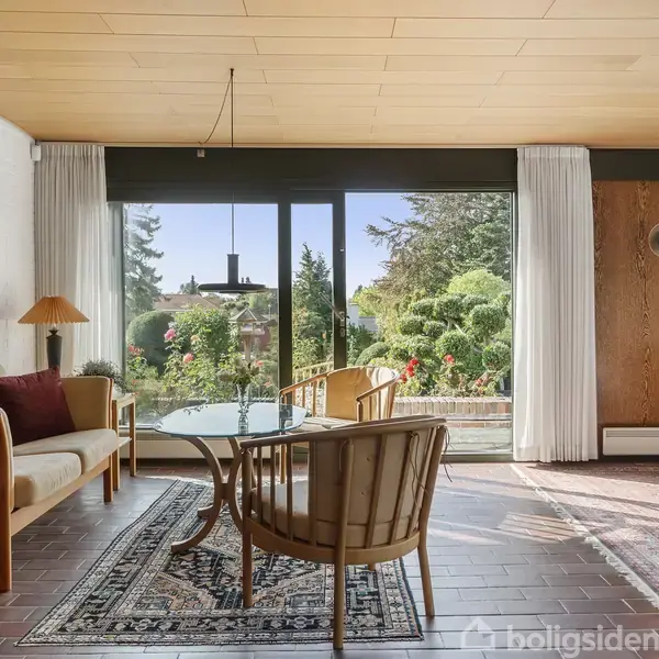 Stue med møbler som sofa og stole omkring et glasbord. Store vinduer giver udsigt til en velplejet have. Rummets design er hyggeligt med tæpper og lamper.