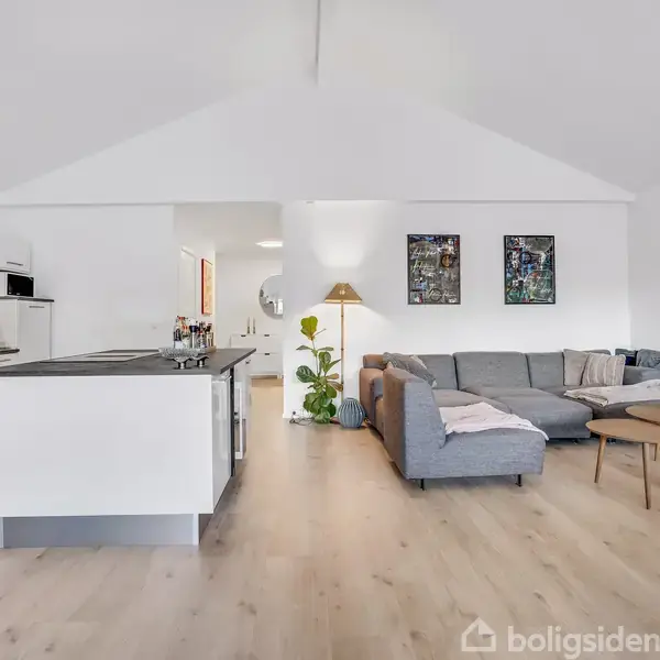En rummelig stue med et moderne køkken til venstre, en stor grå hjørnesofa til højre og kunst på væggen, placeret på et lyst trægulv.