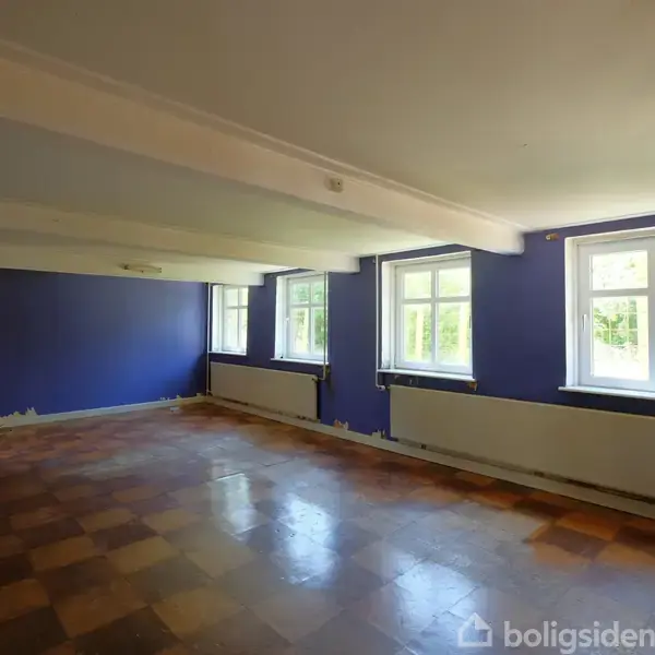 Et tomt rum med mørkeblå vægge og lysebrune ternede gulve, modtager lys fra tre vinduer. Rummet virker rummeligt og enkelt med hvidmalede lofter.