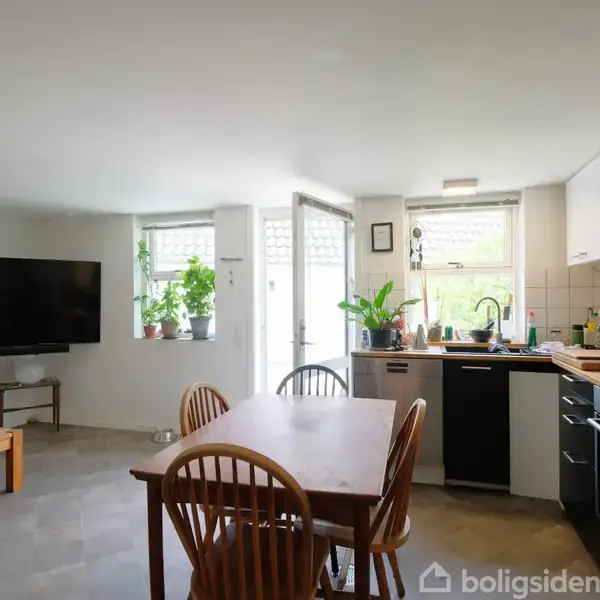 Køkken-alrum med træbord og stole, foran et stort vinduesparti. Køkkenet har hvide vægskabe, sort bordplade, og håndvask. En fladskærm hænger på væggen til venstre. Grønne planter pynter vindueskarmen.