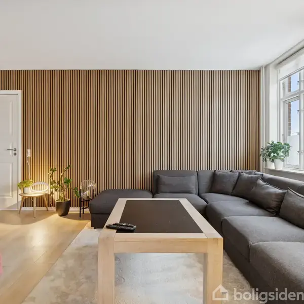 Grå hjørnesofa står i en stue med trævægpaneler, ved siden af store vinduer. Et lyst sofabord med fjernbetjeninger foran, og planter i hjørnet tilføjer et hyggeligt præg.