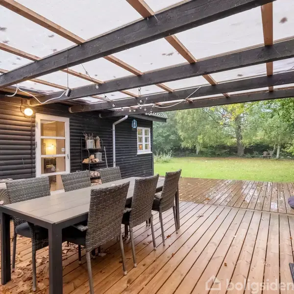 Terrasse med sorte trævægge og gennemsigtigt tag, rummer spisebord med seks stole. Omringet af græsplæne og træer. Indendørs lys er synligt gennem vinduer. Der ligger blade på gulvet.
