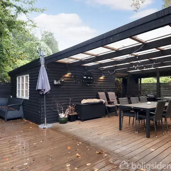 Terrasse med sorte træplanker, havebord og stole, grill i baggrunden. Hyggelig udendørs siddeplads med sofa og parasol. Omgivet af træer og hegn for privatliv.