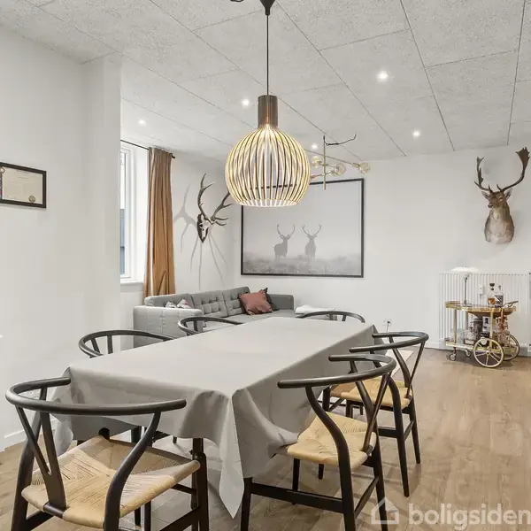 Et spisebord med seks stole er dækket med en dug. En moderne lampe hænger over det. I baggrunden er en stue med sofaer, hjortevægdekorationer og lyse trægulve.