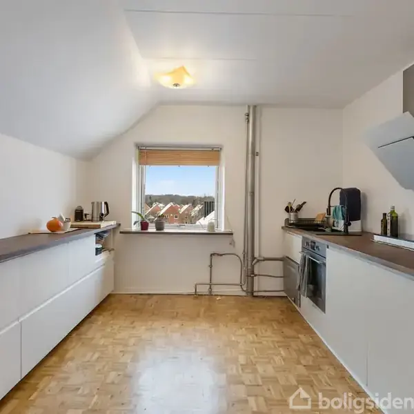 Et køkken med hvide skabe og en ovn indbygget. Bordpladerne er træfarvede. Et vindue viser bytagene udenfor. Køkkenredskaber og en skål ses på bordet.