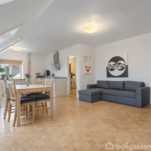 Gråt hjørnesofa står i en lys stue ved siden af et spisebord med stole. Køkken ses i baggrunden med planter og dekorationer. kunstværk på væggen viser tre ansigter.