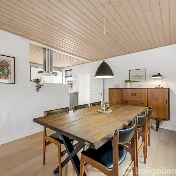 Et spisebord med seks stole står i et lyst, moderne rum. Over bordet hænger en sort lampe. I baggrunden ses et skab, vægkunst og en delvis synlig køkkensektion.
