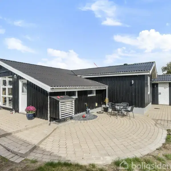 Sortmalet træhus med store vinduer står på en flisebelagt terrasse. Der er havemøbler og blomsterkrukker i en solrig have under en blå himmel.