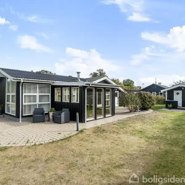 Sort sommerhus står stille på græsplæne med flisebelagt gangsti rundt, omgivet af hæk og buske under blå himmel. I baggrunden ses et mindre skur.