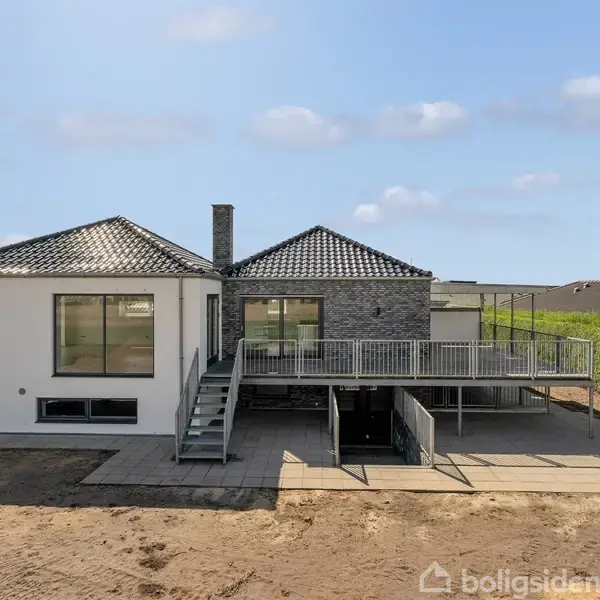 Et moderne hus med sort tegltag har en stor terrasse med trapper ned til en gårdsplads. Omkring huset er der nyplantet græs, og baggrunden viser en høj hæk.