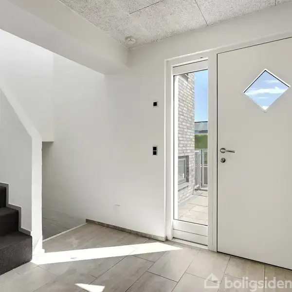 Trappe med mørkt tæppe fører opad ved siden af en hvid dør med firkantet vindue. En lys gang med flisegulv og glasdør ud mod en terrasse. Ingen tekst.