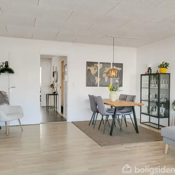 Spisestue med et træbord, omkranset af grå stole. En glaskabinet står ved siden af. Rummet har lyse trægulve, hvidmalede vægge, en grå sofa og elegant belysning.
