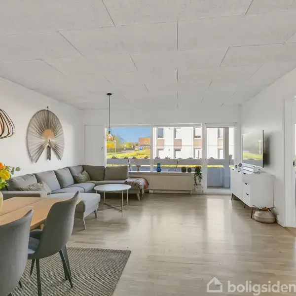 Stue med grå sofa og træbord, dekoreret med blomster, placeret ved store vinduer. Spisebord med stole og lampe hængende over. TV på væggen. Indgang til andet rum.