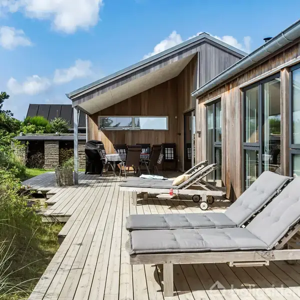 To liggestole står på en træterrasse foran et moderne træhus, omgivet af grønne planter og blå himmel. I baggrunden ses en siddemøbelgruppe under en overdækket sektion.