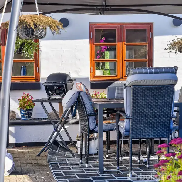 Bord og stole står under en pavillon på en terrasse. Omkring er blomsterkrukker, en grill og et hvidt hus med brune vinduer. Hængende planter skaber en hyggelig atmosfære.