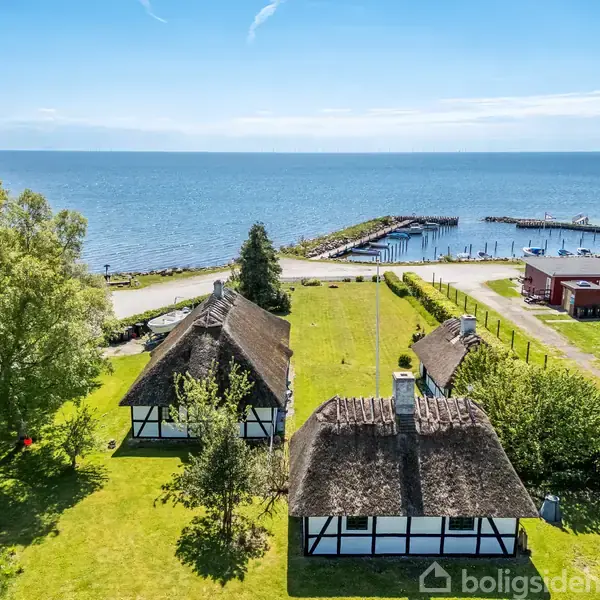 To bindingsværkshuse med stråtage ligger i en velplejet have ved siden af en kyst. I baggrunden ses en mole og bådehavn, omgivet af træer og grønt græs.