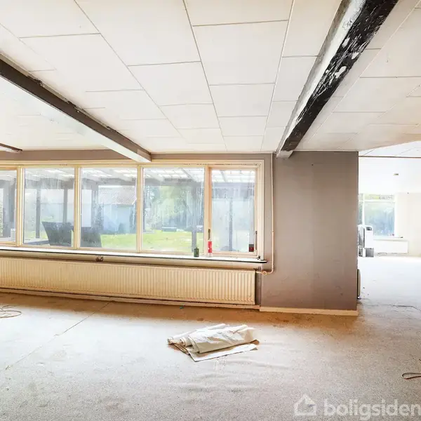 Et tomt rum med store vinduer indrammer et syn ud til en grøn have. Træbjælker løber hen over loftet, og gulvet ser ud til at være under renovering.