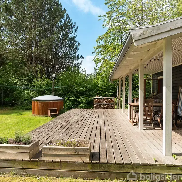 Træterrasse med havemøbler og en brændestabel på en skyggefuld veranda. En grønt græsplæne og et trækar ses i baghaven omkranset af frodig vegetation.