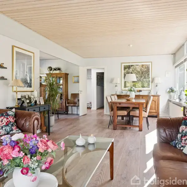 Stue med brune lædersofaer dekoreret med blomstrede puder, står rundt om et glasbord med en vase med blomster. I baggrunden ses et spisebord og store vinduer med planter.