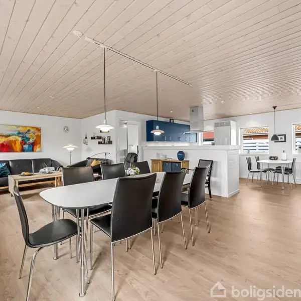 Stue med spisestue og køkken i åben planløsning. Spisebord med seks stole i forgrunden. Moderne møbler og farverig kunst på væggen. Lyse trægulve og lofter.