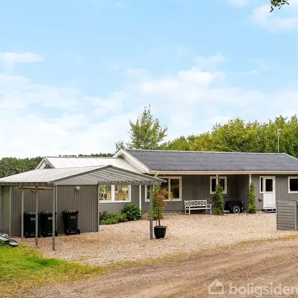 Et gråt træhus står stille. Det har en veranda med havemøbler, omgivet af en græsplæne og skovklædte områder med høje træer i baggrunden.