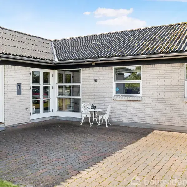 En hvid murstensbungalow med store vinduer står ved en flisebelagt gård. Et lille havesæt med to stole og et bord ses ved indgangen under en klar blå himmel.