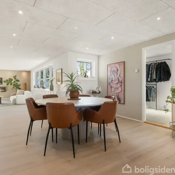 En spisestue med runde borde og brune stole står i et lyst rum med trægulve. Til venstre er en stue med sofaer, og til højre en åben garderobe.