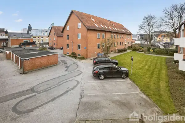 Rødt murstensbyggeri står stille på en parkeringsplads. To biler holder parkeret ved siden af græsplæne. Omkring ses bymiljø og flere træer.