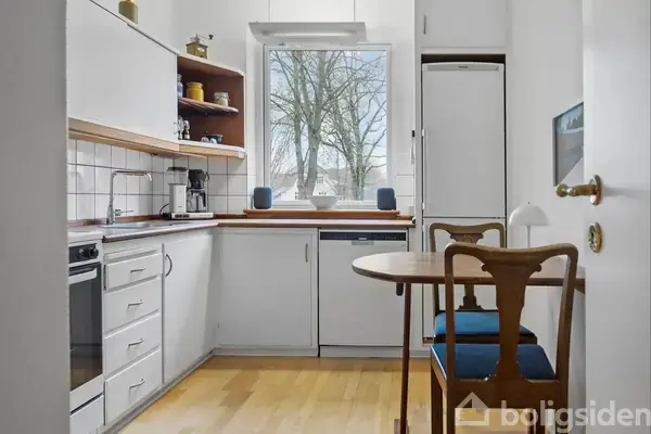 Et kompakt køkken med hvide skabe og træbordplade. Rundt bord med to stole står ved vinduet, som viser et træ udenfor. Trægulv giver en varm atmosfære.