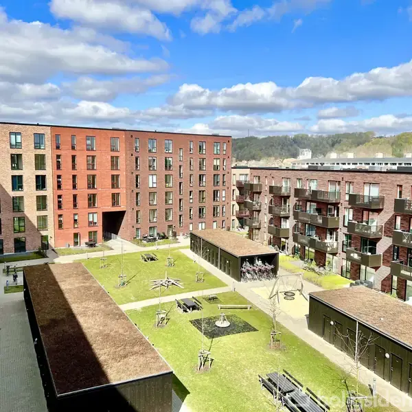 Et moderne boligkompleks med flere etager omgiver en gårdsplads med grønne områder, træer og bænke. Solen skinner på bygningerne, og himlen er let skyet.