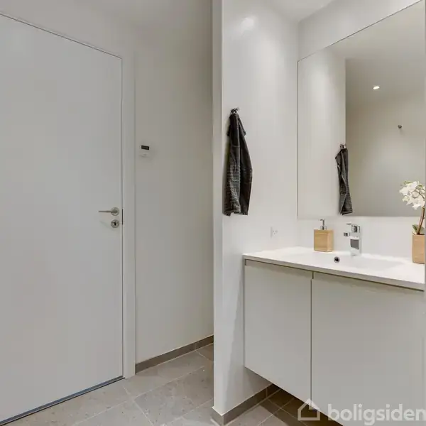 Badeværelse med vask på hvidt skabsmodul, spejl over vasken. En sort håndklæde hænger på væggen. Dør til venstre, beige gulvfliser. Blomst i vase på vask.