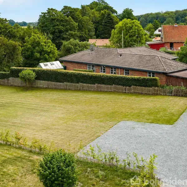 En enkel bygning med fladt tag er omgivet af en velplejet græsplæne og hæk. Bagved ses træer og forreste er en grusvej. Huse anes i baggrunden blandt grønne områder.