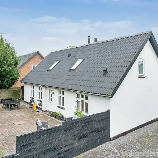 Hvidt hus med sort tag står på en brostensbelagt terrasse, omgivet af grøn have og træhegn, i et boligområde med flere huse i nærheden.