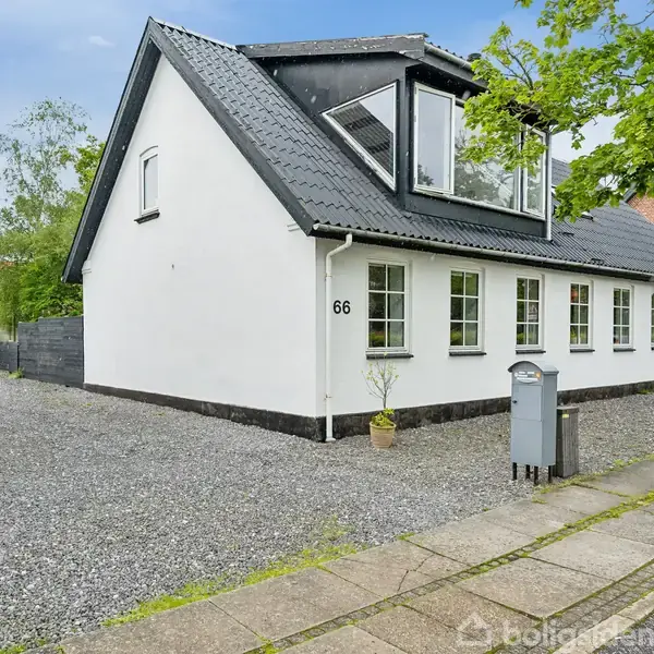 Hvidt hus står stille langs en grusbelagt indkørsel, omkranset af en grøn have og fortov. Husnummeret "66" ses tydeligt.
