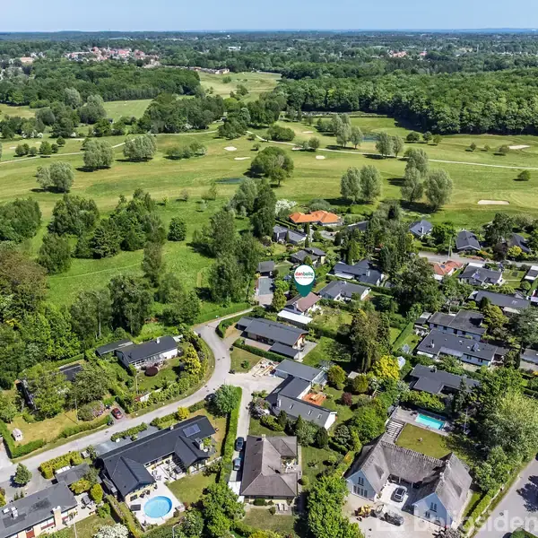 Boliger ligger langs snirklede veje omgivet af træer og haver, mens en golfbane med grønne fairways og træer strækker sig i baggrunden under en klar, blå himmel.
