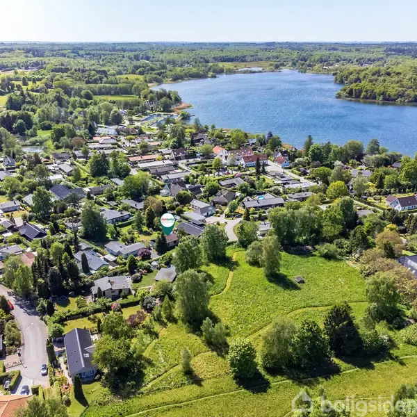 Husområdet strækker sig mod en stor sø omgivet af frodige skove og marker, der skaber et idyllisk landskab. Boliger er spredt langs grønne veje, indrammet af tæt vegetation.