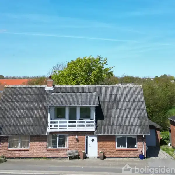 Et rødstenshus med sort tag står langs en vej. Det har en hvid dør og vinduer. En garage ligger til højre. Landskabet er grønt med træer i baggrunden.