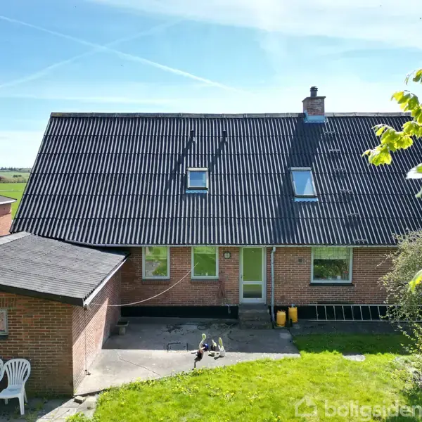 Et rødstenshus med sort tag har to ovenlysvinduer. Huset står på en græsplæne med terrasse og træstole. Der er nogle redskaber langs muren, og landskabet er grønt og åbent.