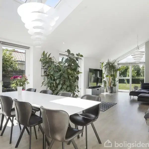 Et langt spisebord med sorte stole står i et lyst rum. Store vinduer og grønne planter skaber en indbydende atmosfære. I baggrunden ses en stue med sofaer og et TV.