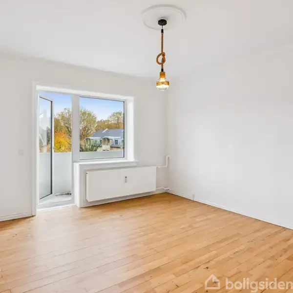 Et tomt rum med trægulv og hvidmalede vægge. En enkelt loftslampe hænger fra midten. Et stort vindue og dør fører ud til en lille altan med udsigt over træer.