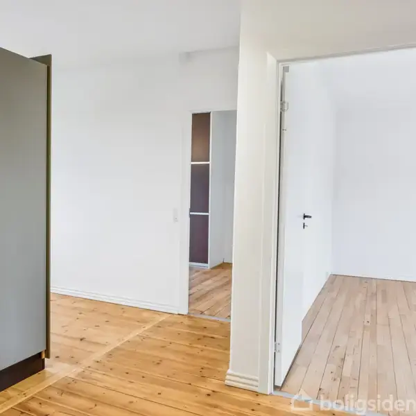 Et lyst rum med trægulv; en åben dør leder til et tilstødende værelse. Der er et skab med mørke låger til venstre i billedet.