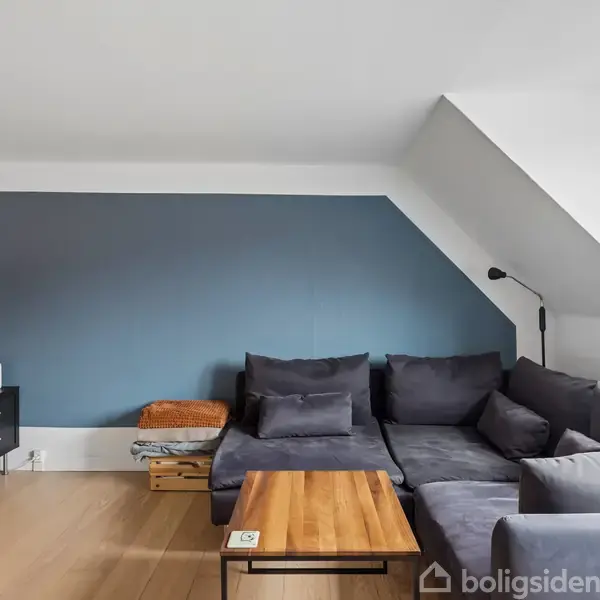 En grå sofa står i en stue mod en blå væg. Et firkantet træbord står foran sofaen. En væglampe og tv er synlige, med vindue til højre.