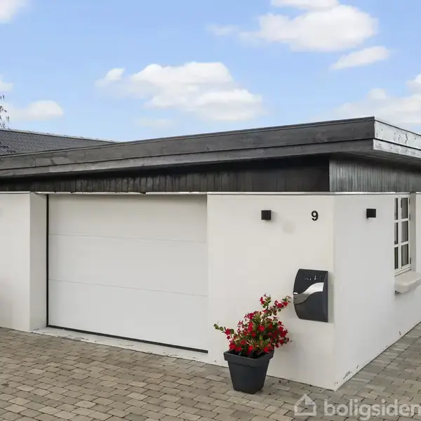 En hvid garage med sort tag har en lukket port. Nummer 9 er på facaden. To blomsterpotter står foran indgangen. Omkring er der en flisebelagt vej og en græsplæne.